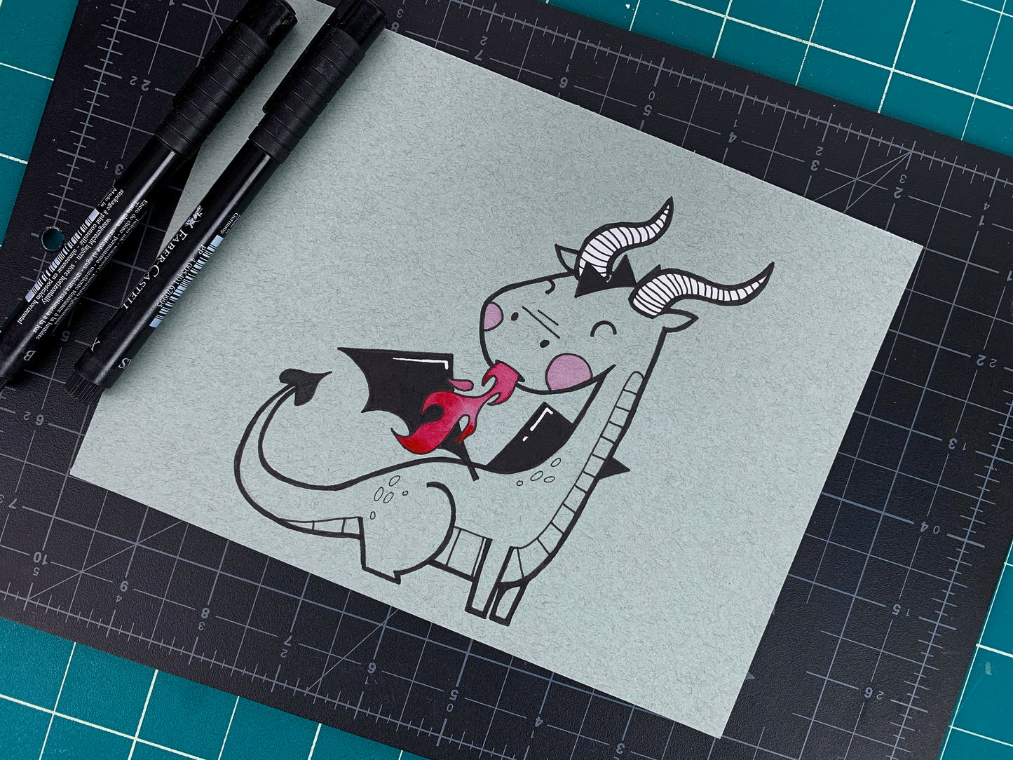 Studio shot of an original illustration of a cute dragon with pink cheeks, horns, and wings that is breathing fire. Made using ink, watercolor, and gouache.