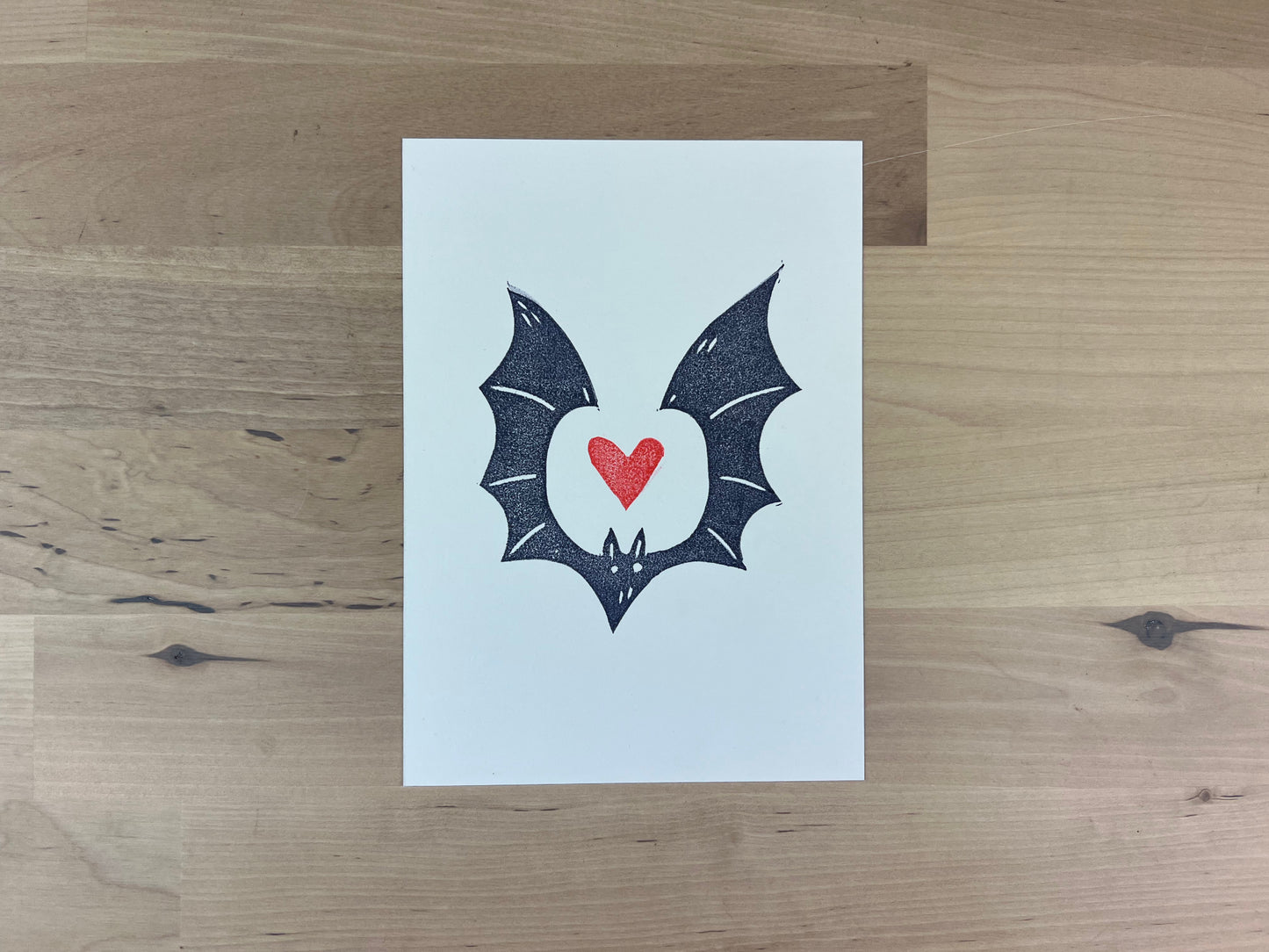 Hand printed illustration of a navy blue bat with a red heart between it's wings.