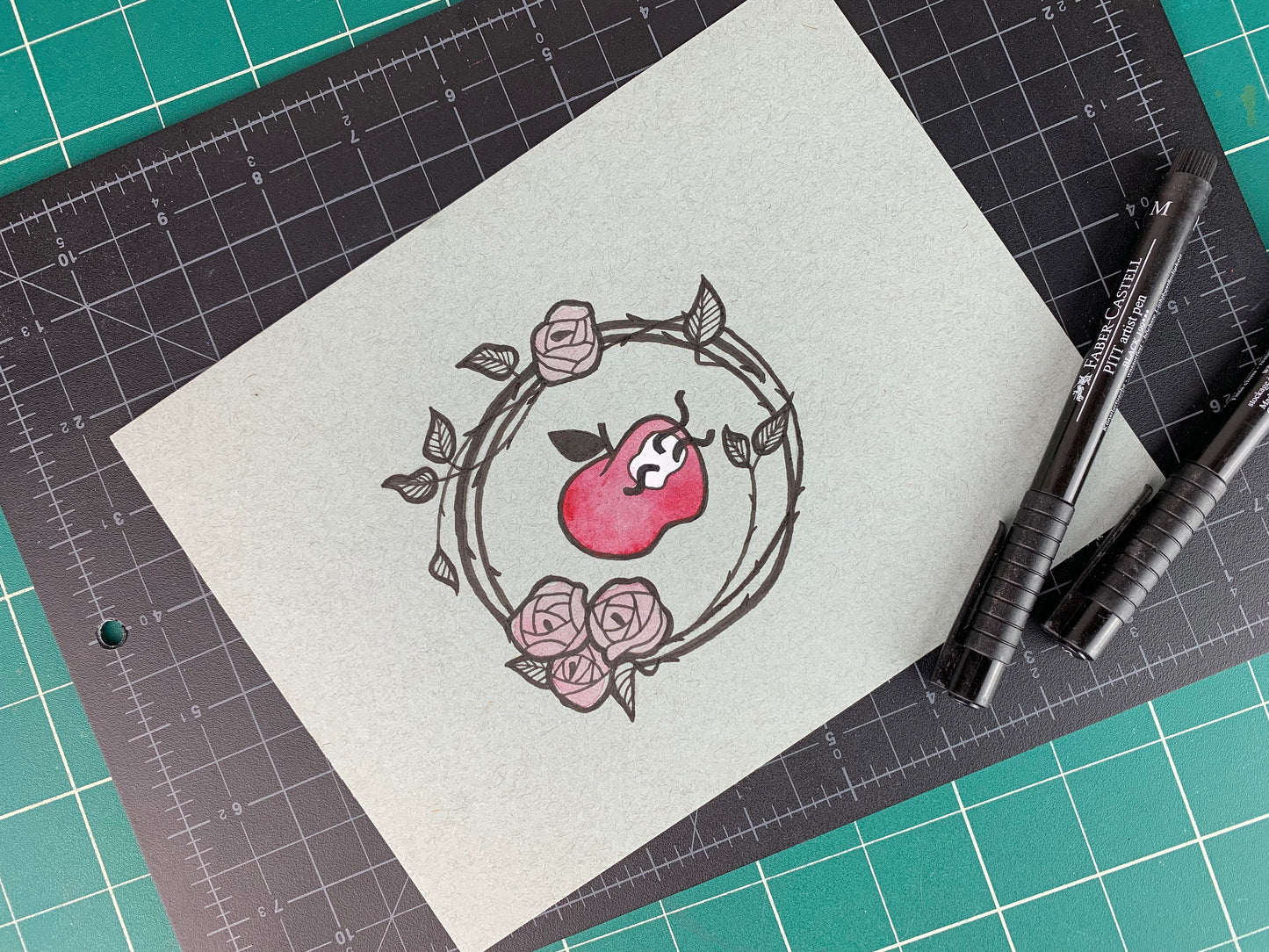 Studio shot of an original illustration of an apple with three little worms coming out of it that is surrounded by a wreath of rose vines. Made using ink, watercolor, and gouache.