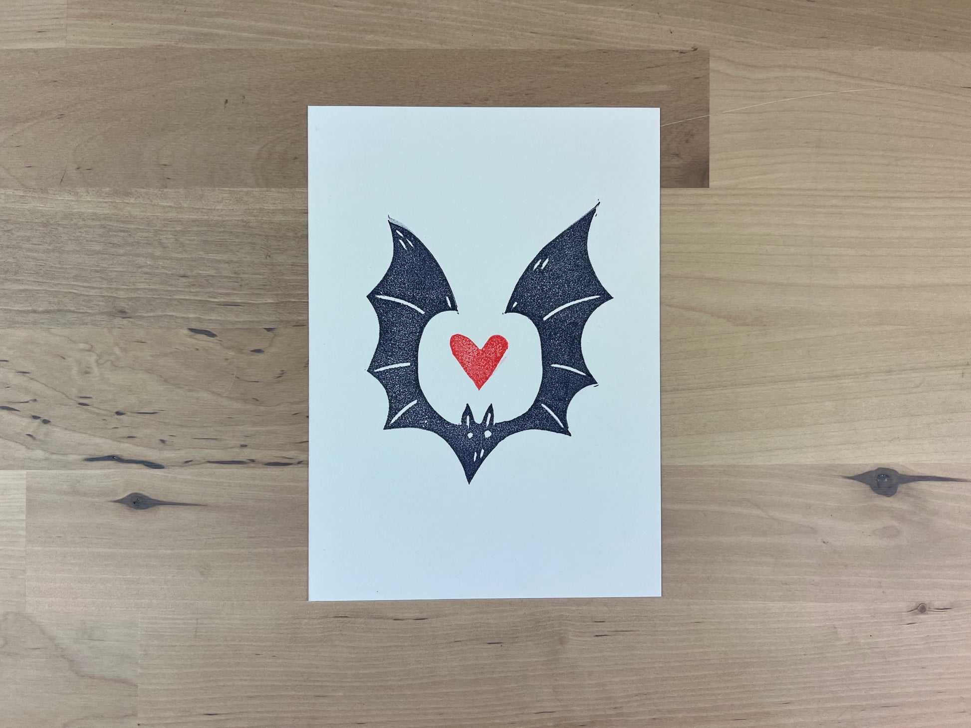 Hand printed illustration of a navy blue bat with a red heart between it's wings.