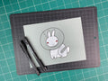 Studio shot of an original illustration of a white rabbit (bunny) wearing a cone collar (cone of shame) with a band-aid over an injurred leg. Made using ink, watercolor, and gouache.