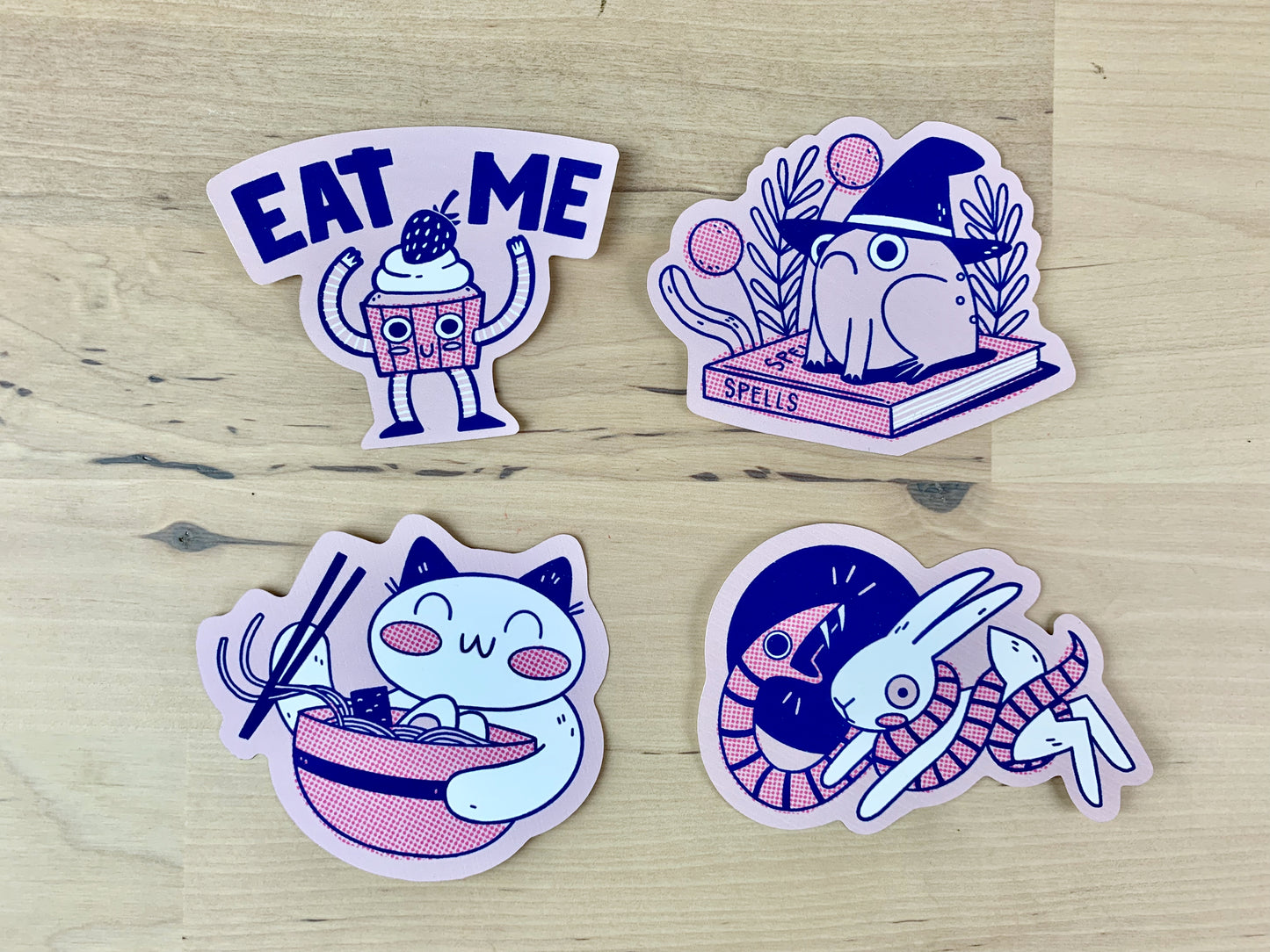 Collection of stickers: Anthropomorphic cupcake with the words "Eat me" above it, a frog wearing a witches hat sitting on top of a book of spells, a lucky cat eating ramen with chopsticks, and a snake wrapping itself around a scared hare/rabbit.