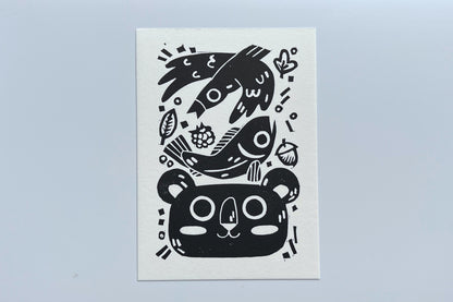 Black ink block print of a bird, fish, leaves, berry, acorn, and bear.
