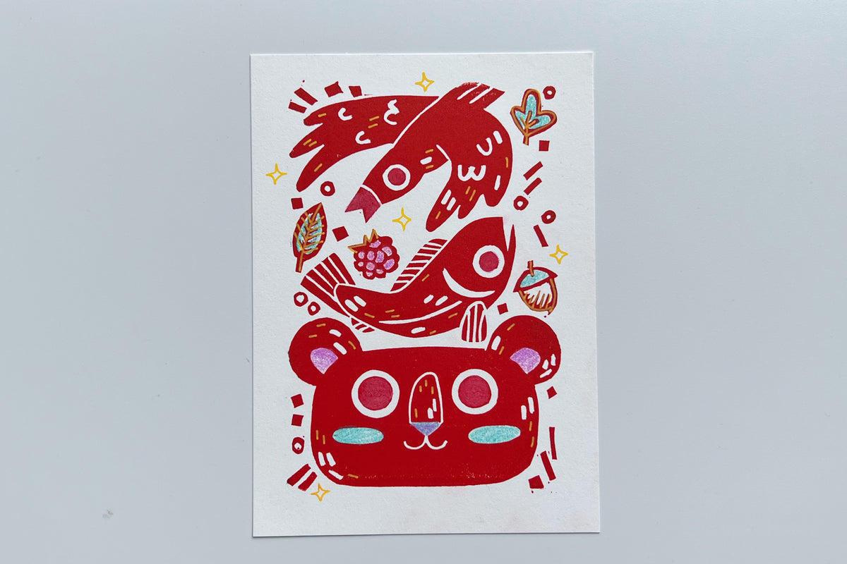 Red ink block print of a bird, fish, leaves, berry, acorn, and bear with colored pencil and POSCA pen coloring.
