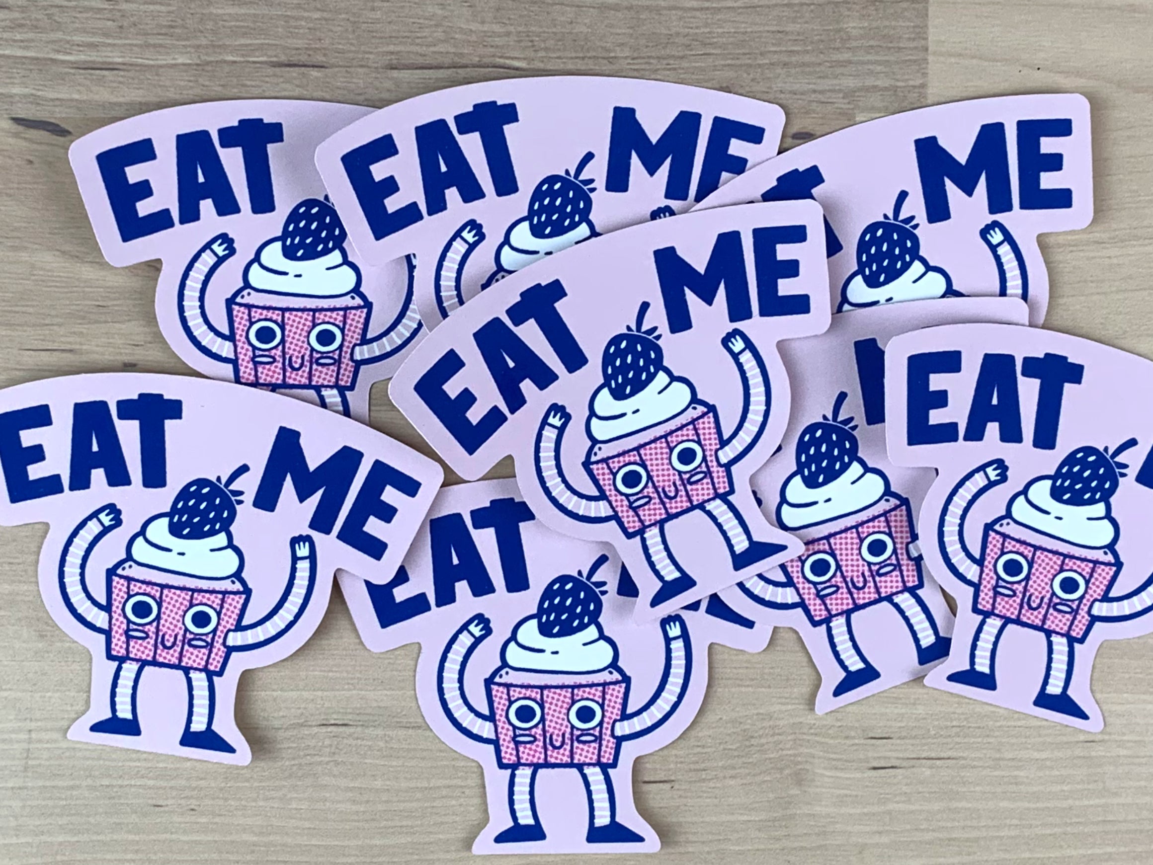 Stickers in pink and blue of an anthropomorphic cupcake with the words "Eat me" above it.