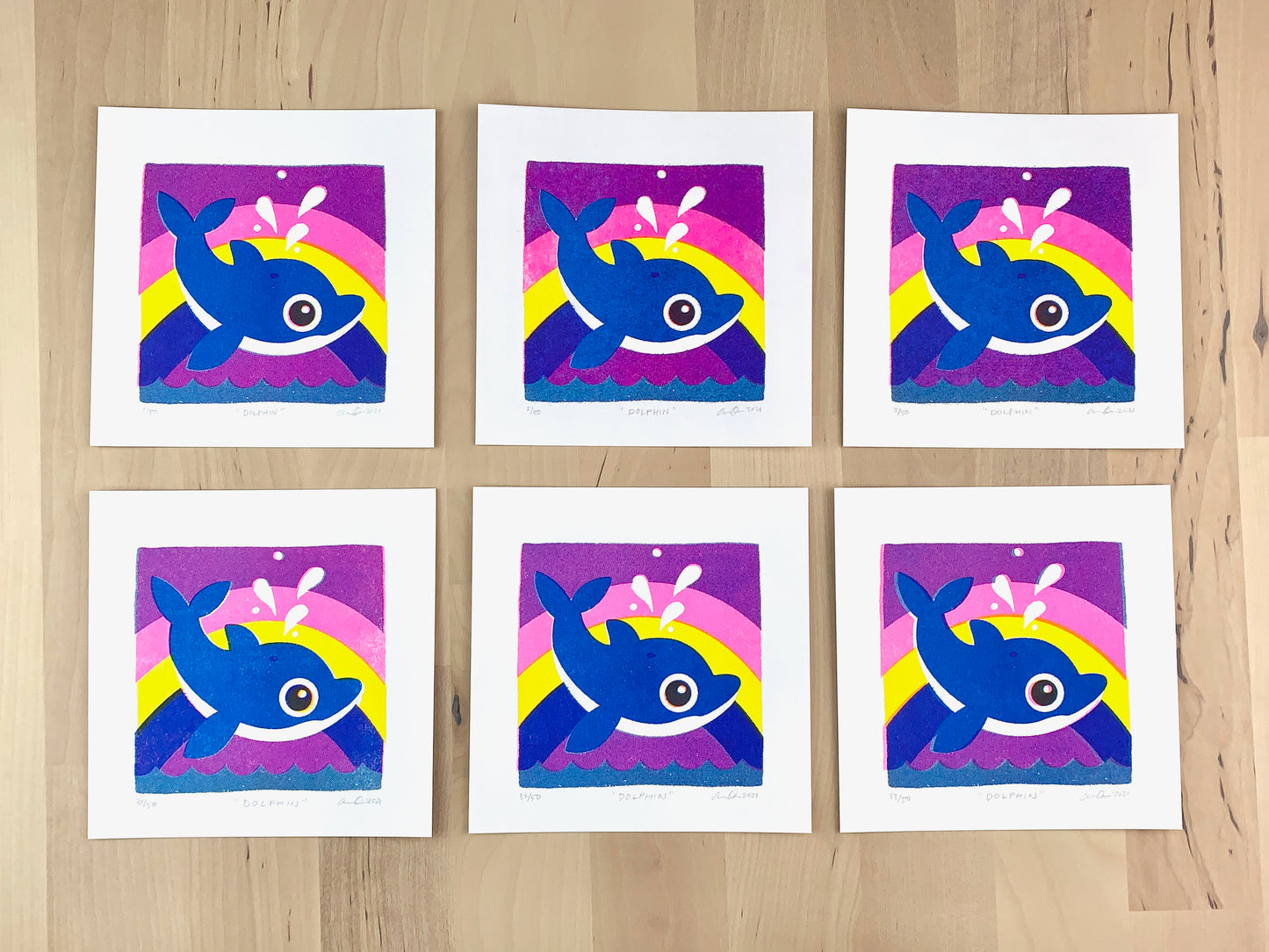 Six Risograph prints of an illustration of a cute dolphin jumping out of the water with a rainbow behind it showing the unique variations.