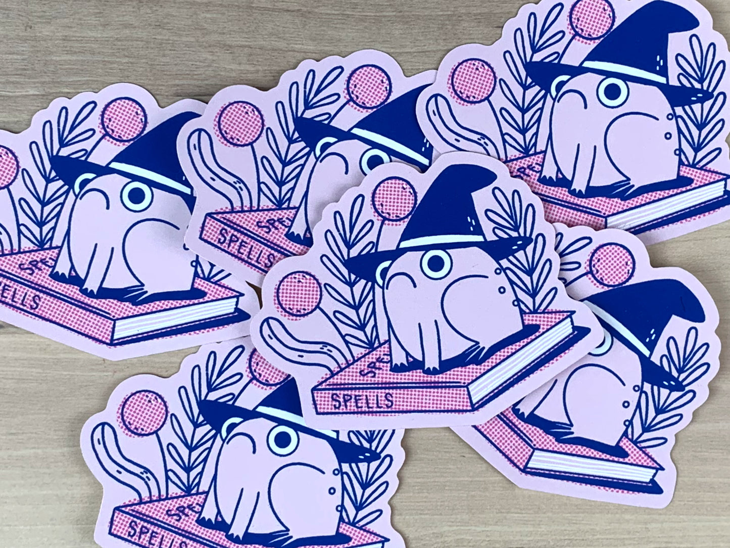Stickers in pink and blue of a frog wearing a witches hat sitting on top of a book of spells.