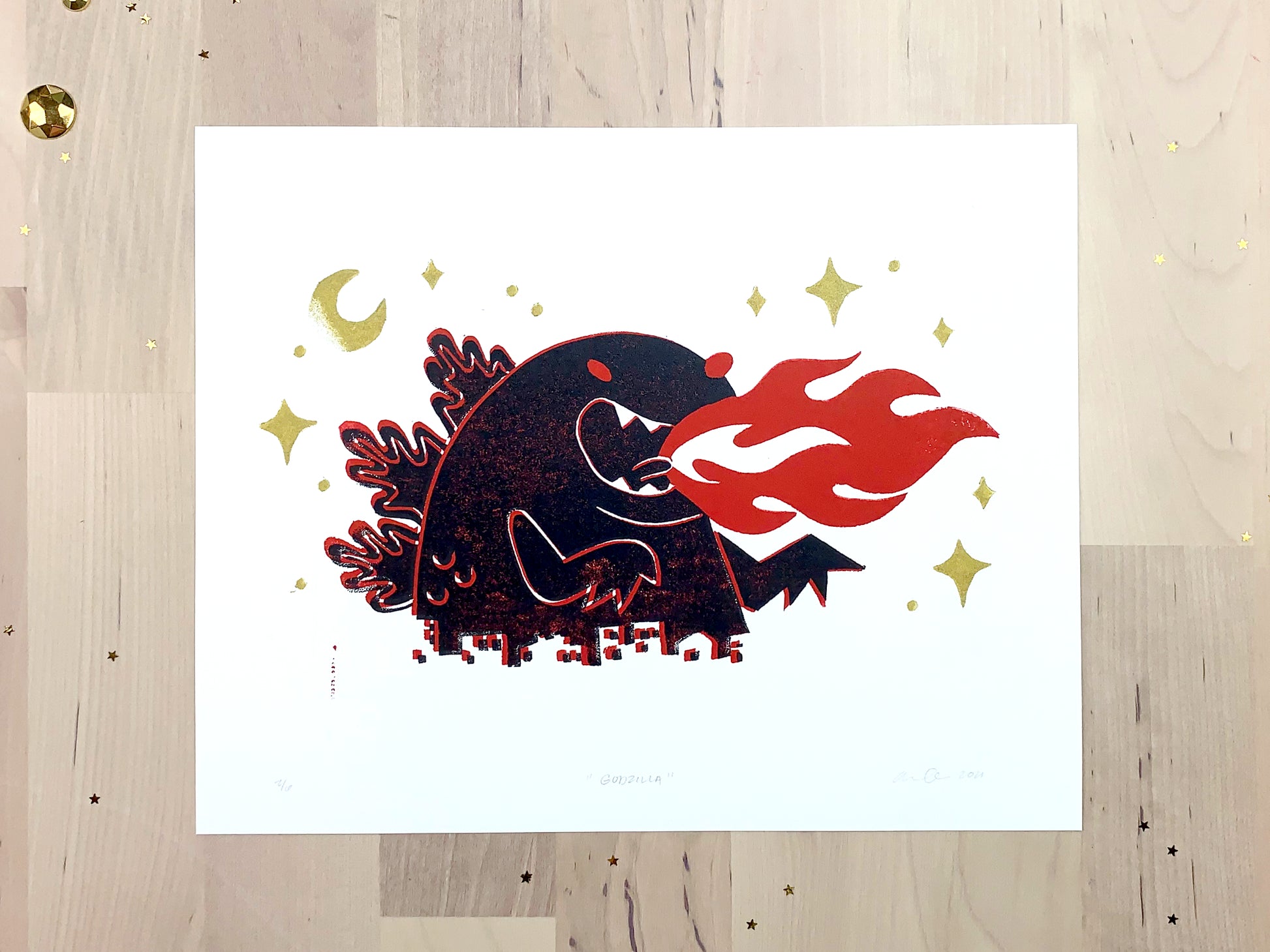 Original limited edition #2 art print by Amber Orenstein. Two color, red and black, reduction block print of a Japanese kaiju monster roaring with flames while destroying a city with gold stars and moon.