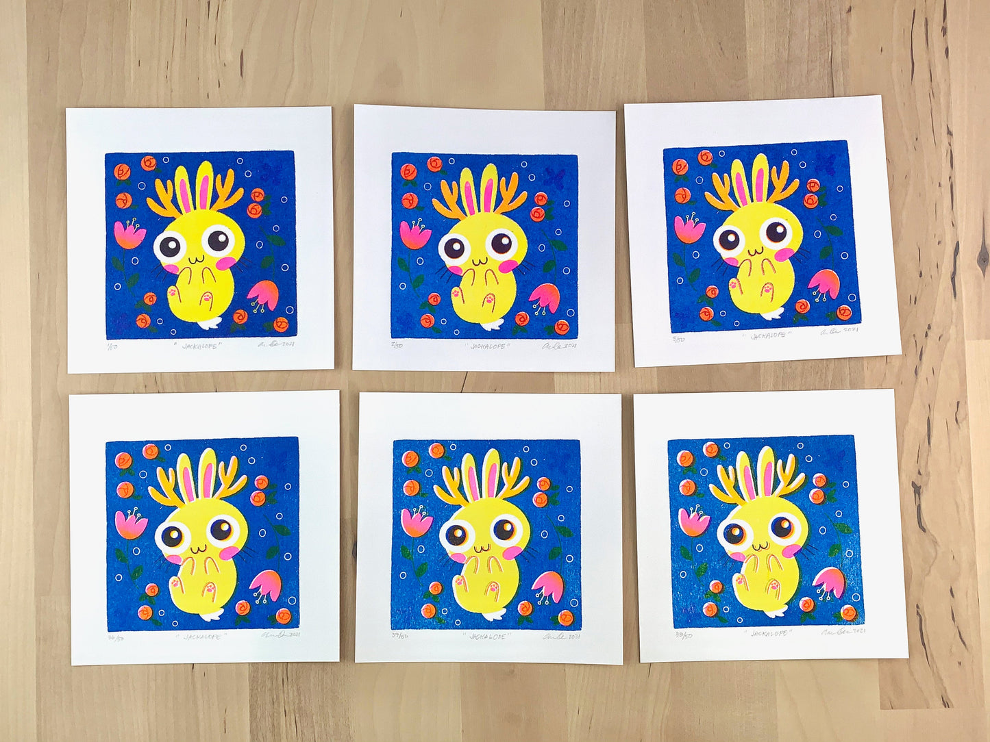 Six Risograph prints of a cute yellow jackalope bunny illustration on a blue background and flowers showing the unique variations.