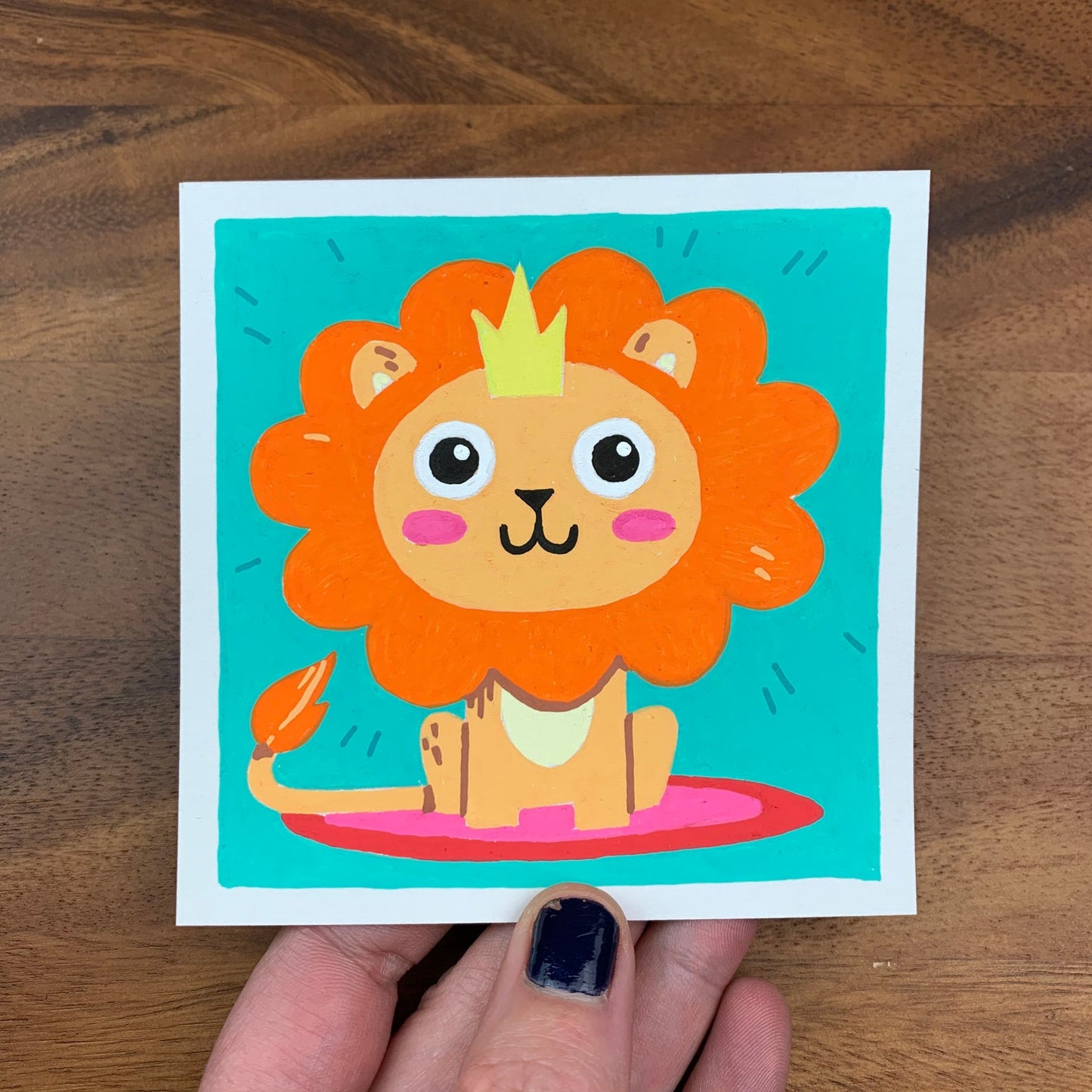 Original artwork of a cute orange lion with a crown sitting on a rug on a teal background. Materials used: Uni-Posca paint markers.