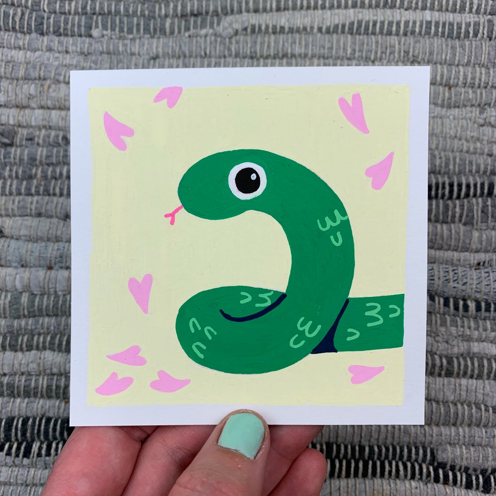 Original artwork of a cute green snake on a light yellow background with some pink petals falling around it. Materials used: Uni-Posca paint markers.