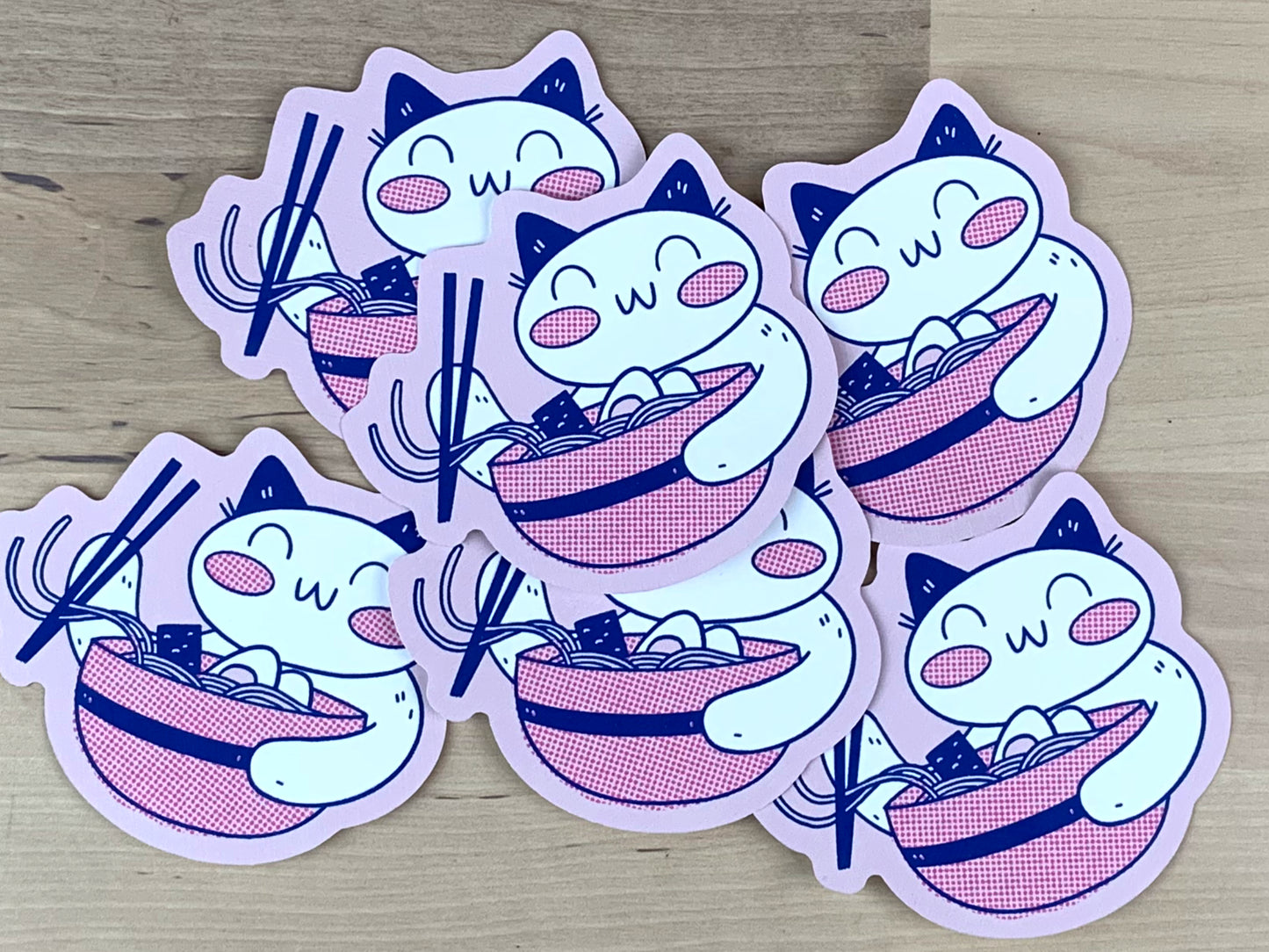 Stickers in pink and blue of a lucky cat eating ramen with chopsticks.
