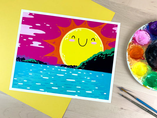 Overhead shot of an artist's palette, paintbrushes, and a print. Artwork depicted in print is a bright colorful illustrative style natural scene of a sun with a happy smiling face setting over the Columbia River in the Pacific Northwest.