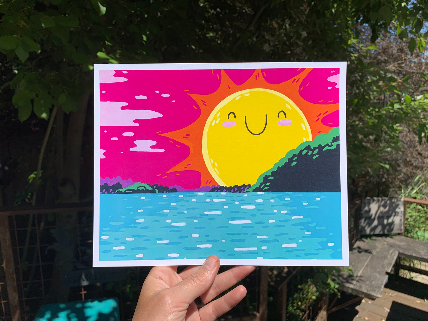 Outdoor shot of a person holding a print. Artwork depicted in print is a bright colorful illustrative style natural scene of a sun with a happy smiling face setting over the Columbia River in the Pacific Northwest.